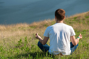 Young man sits on the top of the hill viewing wide Dnister river. Yoga meditation
