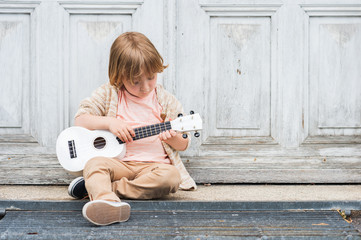 Little happy boy plays his guitar or ukulele, sitting by the wooden door outdoors