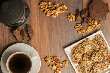 Overhead view of french press coffee, chocolate, nuts, and cookies on a dark rustic table with empty space in the center for text or graphics.