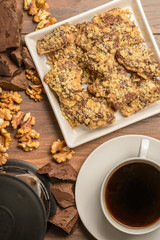 Overhead view of a plate of homemade chocolate toffee walnut cookies with ingredients and a fresh brewed cup of coffee on a rustic wood table.