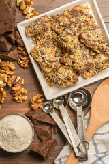 Overhead view of homemade chocolate walnut cookies with ingredients and kitchen utensils on a dark rustic wood table.