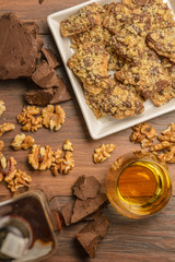 Overhead view of homemade chocolate walnut cookies with ingredients and a bottle and glass of scotch whisky on a dark rustic table.