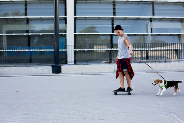 cool young and handsome caucasian brunette hipster skater guy wearing a hat posing smiling and having fun outside while skating with his skateboard in the city with his beagle pupppy dog