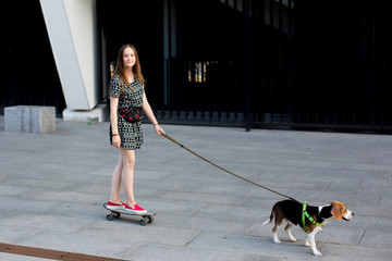 portrait of cool young and beautiful caucasian blonde teenager hipster girl with long gorgeous hair is posing smiling and having fun  skating on a skateboard with her beagle puppy dog