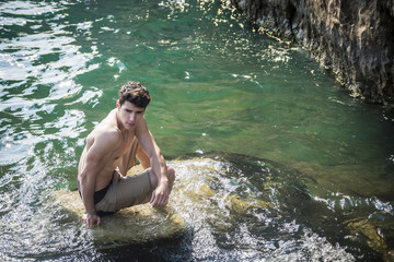 Young shirtless athletic man sitting on rock by sea