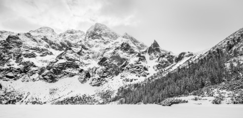 Fototapeta premium Black and white panorama landscape of a frozen snow covered alpine lake and mountain peaks on a cloudy winter day.