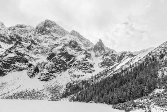 Fototapeta Black and white landscape of a frozen snow covered alpine lake and mountain peaks on a cloudy winter day.