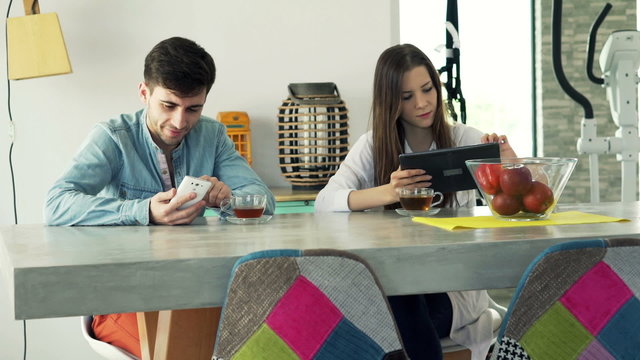 Young couple with smartphone and tablet computer sitting by table at home
