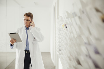 Mature man in lab coat on phone looking at digital tablet