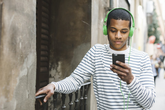 Spain, Barcelona, young man hearing music with green headphones on street