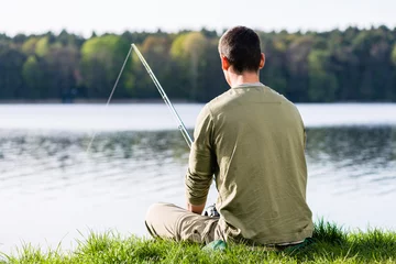 Fototapeten Angler, der im Gras am Seefischen mit seiner Rute sitzt © Kzenon