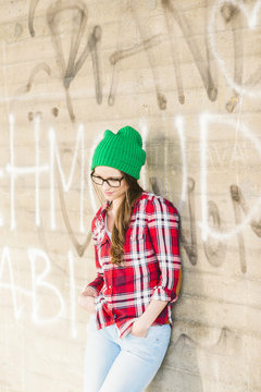 Young woman with checkered shirt and green wooly hat leaning at graffiti wall