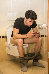 Young Man with Cell Phone Sitting on Toilet