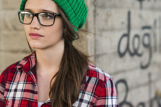 Young woman with checkered shirt and green wooly hat at graffiti wall