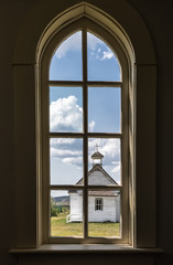 image from inside an old church looking out the arch window at another old church with blue sky in the summer time