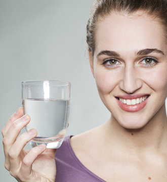 smiling young beautiful woman wearing purple shirt displaying glass of pure tap water