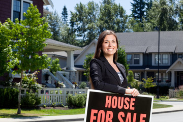Professionally dressed real estate woman outside with a house for sale sign.