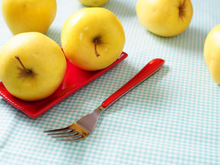 Still life with yellow apples, red plate and red fork on checkered table cloth