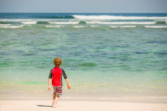 Cute 7 years old boy in red rushwest swimming suit at tropical beach with white sand and green ocean