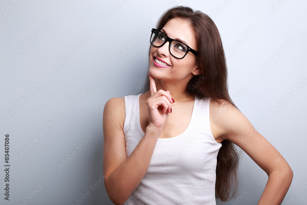 Wall mural Healthy young thinking woman in glasses looking up on blue backg