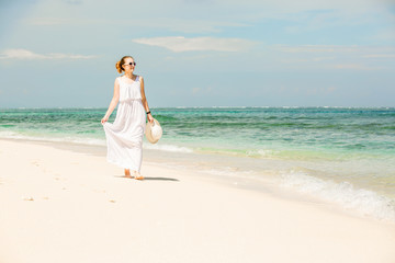 Young woman in long white dress holding hat walks along tropical beach having great summer time on holidays