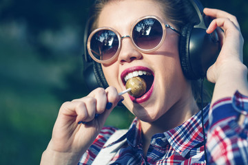 Hipster girl listening to music on headphones with lollipop.