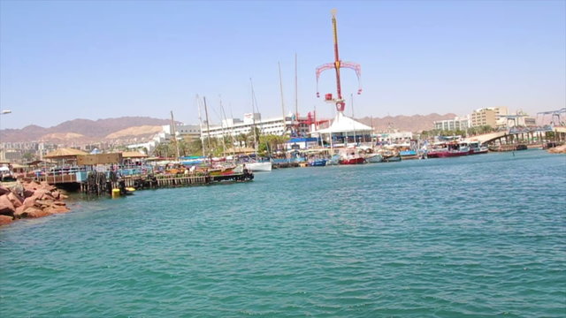 Boat tracking shot of the entrance to the marina of Eilat.
Boats are near the pier. Eilat boardwalk is on the horizon.