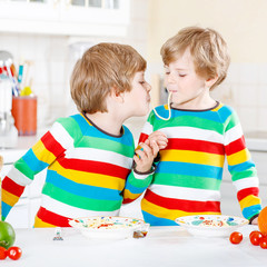 Two little kid boys eating spaghetti in domestic kitchen.