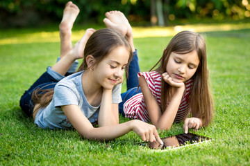 Two sisters lying on grass outdoors and playing on digital table