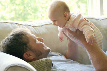 A happy young father is lifting his baby girl up over his head and playing with her as he relaxes at home on the couch.