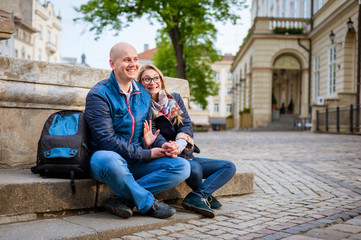 Young fashion elegant stylish couple, travel by old European cities, sitting on an old stone, with a backpack, on the square with paving stones