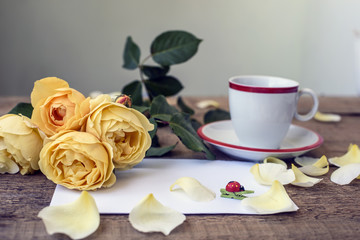 Obraz na płótnie Canvas yellow roses, coffee cup and white sheet of paper on old wooden table