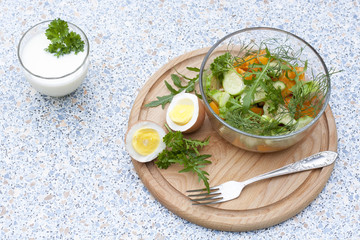 Salad with fresh vegetables and herbs in a glass bowl on a wooden board, boiled eggs and yogurt with herbs