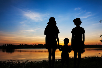 Obraz na płótnie Canvas silhouette three child girl and boy standing hand hold and looking forward at the river sunset background