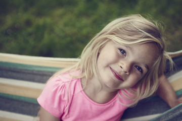 Fototapeta premium Portrait of blond child girl with blue eyes looking at camera and smiling, relaxing on a colorful hammock in a home garden on summer holiday. Vacation lifestyle kid fun activities.