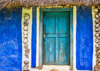 Entrance to an old blue clay thatched roofed house with a turquoise door with cracked paint.