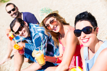 Vier Freunde sitzen mit Getränken am See Strand in der Sommersonne