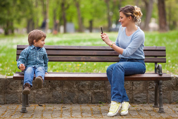 Fototapeta na wymiar Young mother photographing her cute little son sitting on a park bench