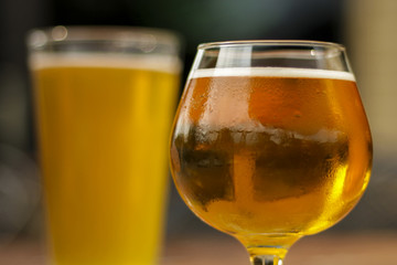 Golden ale beer in two different types of glasses: a hazier lager in a pint glass, and a golden ale in a snifter. The snifter is in focus while the back drink is blurry.