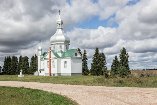 horizontal image of a beautiful white ukrainian church sitting in the country surrounded by spruce trees with a driveway in the foreground under a very cloudy sky in the summer time. 