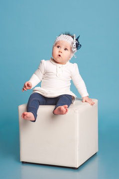 Cute kid girl sitting on white chair in room over blue. Posing in studio. Wearing trendy clothes. Childhood. 
