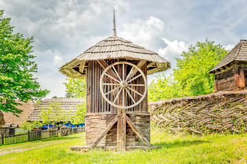 Old wooden water well house with a large wheel near a woven willow fence with green trees in the...
