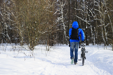 Fototapeta na wymiar Young extreme cyclist with black backpack, in jeans and blue jacket walking with mountain bicycle on snowy track near forest in winter sunny cold day 