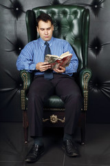 Obraz premium Serious young man reads book in leather armchair in room 