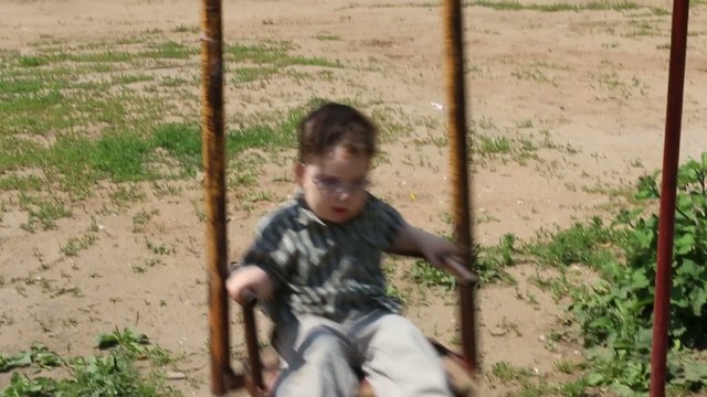 Little cute boy in glasses swings on swing at summer day
