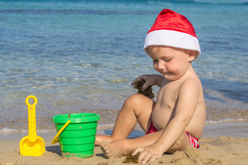 Baby by the sea in a cap of Santa Claus playing with sand