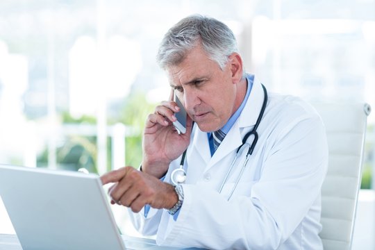 Serious Doctor Working On Laptop And Having Phone Call