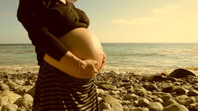 Footage of a pregnant woman on the beach rubbing her bump in sepia/film burn color