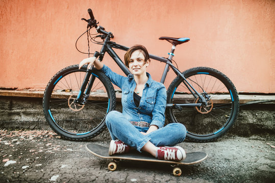 Young sexy brunette girl with short hair standing near vintage bicycle and holding a skateboard, having fun and a good mood while looking in camera and smiling