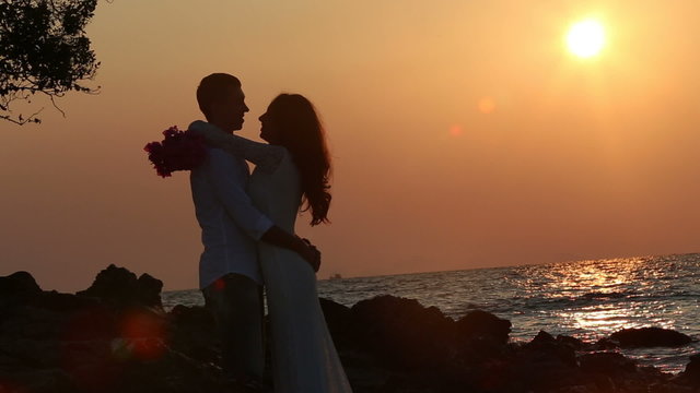 silhouette of bride hugs and kisses groom with bouquet round his neck against sun rising over sea